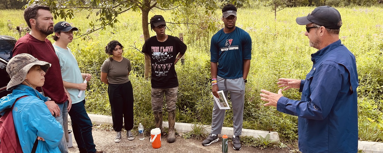 Researchers and interns circle around a male leader with grasses and trees in the background