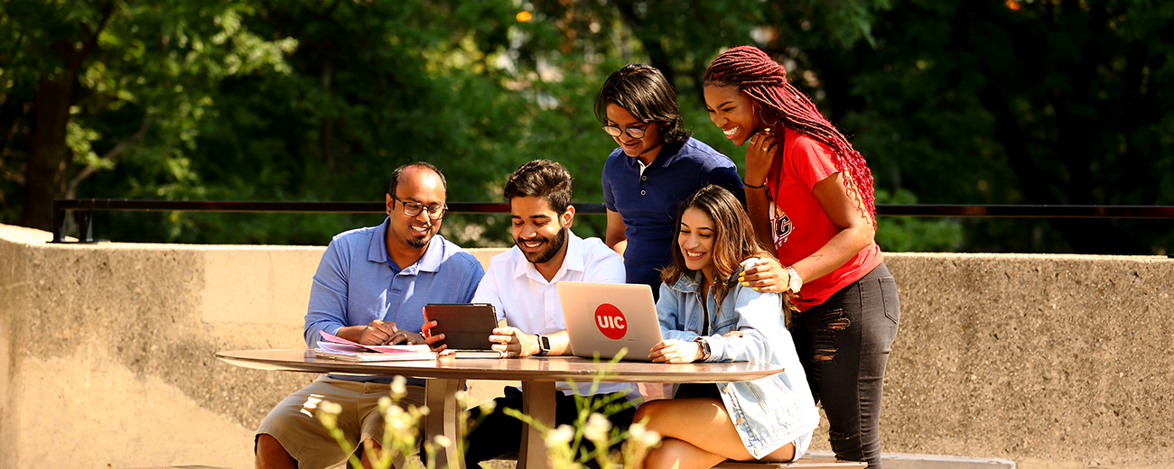 diverse group of students smiling grouped around table looking at UIC laptop