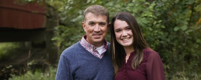 Former high school principal Jason Leahy in outdoor portrait with daughter Emma, tree leaves as background