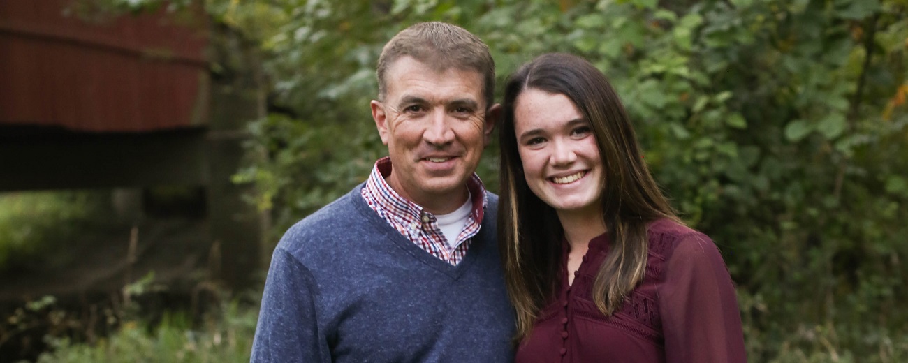 Former high school principal Jason Leahy in outdoor portrait with daughter Emma, tree leaves as background