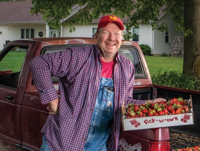 Greg McLaughlin beside a rusty pickup truck in a plaid shirt over overalls and a red ballcap holding a box of strawberries